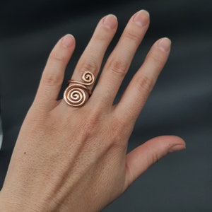 Può includere: Un anello dorato con un design a doppia spirale. L'anello è indossato sull'indice di una mano.