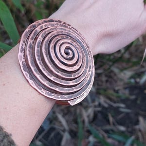 May include: A copper spiral bracelet with a hammered finish. The bracelet is made of multiple concentric circles that create a swirling pattern.