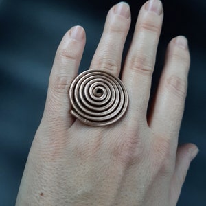 Könnte beinhalten: Ein kupferfarbener Ring mit einem Spiraldesign. Das kreisförmige Design des Rings weist mehrere konzentrische Spulen auf, wodurch ein optisch auffälliges Schmuckstück entsteht. Der Ring wird an einem Finger getragen.