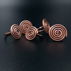Può includere: Cinque anelli di filo di rame con un design a spirale. Gli anelli sono disposti in un gruppo su una superficie nera.