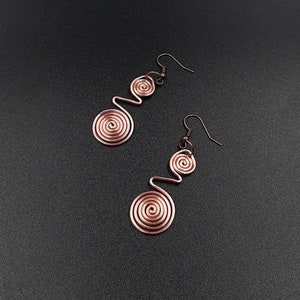 Puede incluir: Un par de pendientes de alambre de cobre con un diseño en espiral. Cada pendiente tiene dos espirales, una más grande que la otra.