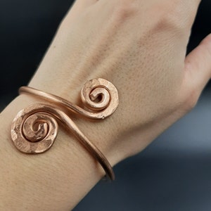 Könnte beinhalten: Ein Kupferarmband mit zwei Spiralmotiven. Das Armband befindet sich an einem Handgelenk.