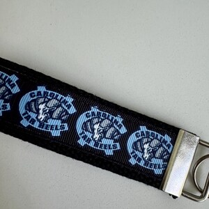 May include: Black lanyard featuring the Carolina Tar Heels logo in light blue and white. The logo is repeated across the lanyard. A silver-colored metal clasp is attached to the end.