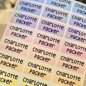 Op de afbeelding: Een vel met rechthoekige zelfklevende etiketten met een holografische afwerking die varieert tussen blauw, roze, oranje en geel. Elk etiket is bedrukt met de naam Charlotte Packer in een vet, zwart en afgerond lettertype.