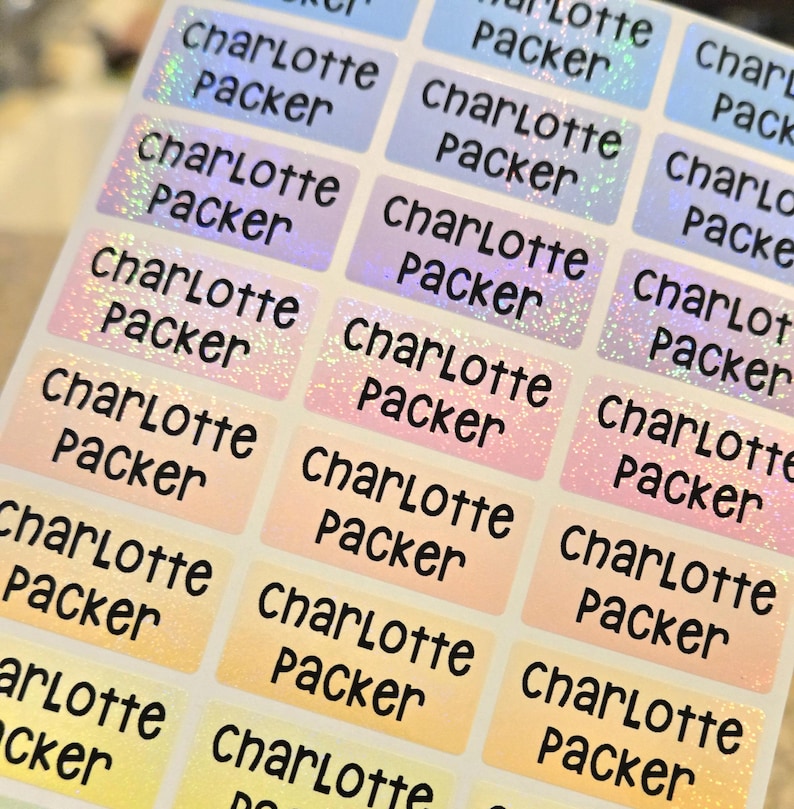 Op de afbeelding: Een vel met rechthoekige zelfklevende etiketten met een holografische afwerking die varieert tussen blauw, roze, oranje en geel. Elk etiket is bedrukt met de naam Charlotte Packer in een vet, zwart en afgerond lettertype.