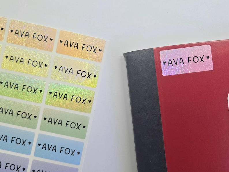 Op de afbeelding: Een vel met rechthoekige zelfklevende etiketten met een holografische afwerking in diverse pastelkleuren, elk bedrukt met de naam AVA FOX geflankeerd door twee kleine zwarte hartjes. E&eacute;n identiek etiket is geplakt op een donkerrood notitieboek.