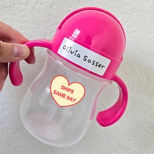 May include: A clear plastic sippy cup with a bright pink lid and two matching side handles. The lid features a white rectangular label with the name Olivia Sasser printed in black text. A heart-shaped sticker is attached to the cup body.