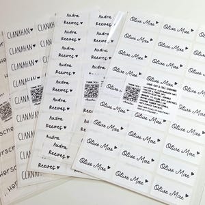 May include: Multiple sheets of white rectangular adhesive labels featuring black printed names including Herschel, Clanahan, Audra Reeves, and Olive Mae, each accompanied by a small heart icon. Small square QR code stickers are also included.