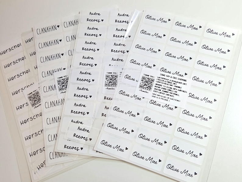 May include: Multiple sheets of white rectangular adhesive labels featuring black printed names including Herschel, Clanahan, Audra Reeves, and Olive Mae, each accompanied by a small heart icon. Small square QR code stickers are also included.
