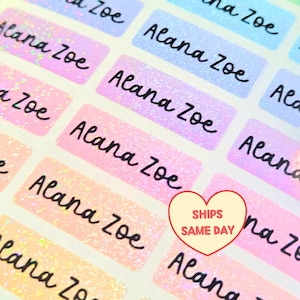 May include: A sheet of rectangular adhesive labels with a holographic glitter finish in pastel shades of pink, blue, and orange. Each label features the name Alana Zoe printed in a black cursive script font arranged in a repeating grid pattern.