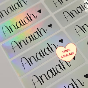 May include: A sheet of rectangular adhesive labels featuring the name Anaiah in a black cursive script, accompanied by a small black heart icon. The labels have a reflective, holographic finish that shifts colors under light, arranged in rows.