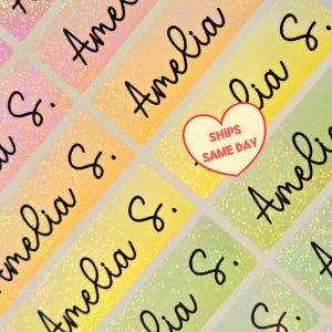 May include: A sheet of rectangular adhesive labels with a glittery, iridescent gradient finish transitioning from pink to yellow and green. Each label features the name Amelia S. printed in black cursive script. A red heart graphic contains the text SHIPS SAME DAY.