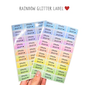 Op de afbeelding: Twee vellen met rechthoekige regenboog glitterlabels met een kleurverloop van blauw naar groen. Elke sticker is bedrukt met de zwarte tekst omonii Short gevolgd door een klein hartje. De tekst bovenaan luidt RAINBOW GLITTER LABEL.