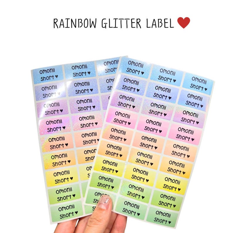 Op de afbeelding: Twee vellen met rechthoekige regenboog glitterlabels met een kleurverloop van blauw naar groen. Elke sticker is bedrukt met de zwarte tekst omonii Short gevolgd door een klein hartje. De tekst bovenaan luidt RAINBOW GLITTER LABEL.