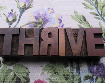 May include: Wooden block letters spelling "THRIVE" in a dark brown hue. The letters are set against a floral patterned background with purple, green, and red flowers.
