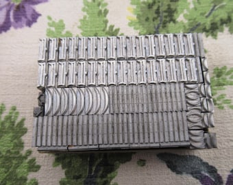 May include: A collection of vintage metal letterpress printing blocks. The blocks are arranged in a rectangular shape, with various letter and design elements visible. The metal has a worn, aged appearance, suggesting historical use.