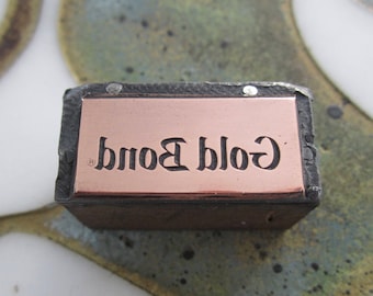 May include: A rectangular metal stamp with the words "Gold Bond" in a stylized font. The stamp has a copper-coloured plate with the text and is set in a dark, textured frame.