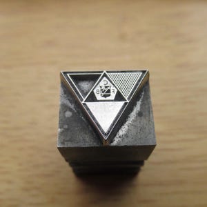 May include: A vintage metal printing block with a triangular design. The design features a triangle within a triangle, with a textured pattern and the letters "C", "B", and "E". The block is a dark gray color.