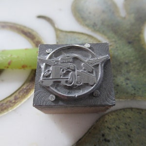 May include: A vintage metal printing block featuring a detailed illustration of a classic truck with wings, set within a circular frame. The block is square, with a wooden base and metal top, and is likely used for letterpress printing.