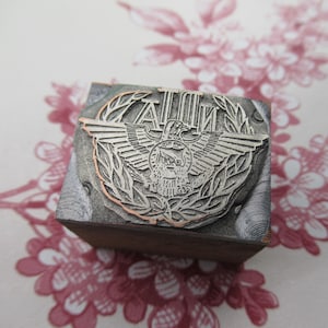 May include: A metal printing block with a design of an eagle with wings spread, surrounded by a wreath. The text "A.D.I.N." is above the eagle.