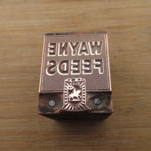 May include: A copper-colored printing block with the text "WAYNE FEEDS 2033" in reverse. The block features a decorative emblem of a horse and rider. The block is set on a wooden surface.