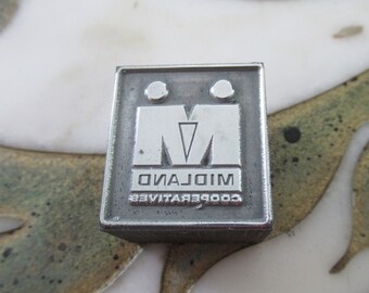 Könnte beinhalten: Ein silberner Metallstempel mit dem Text "COOPERATIVE MIDLAND" und einem stilisierten "M" mit zwei Kreisen darüber.