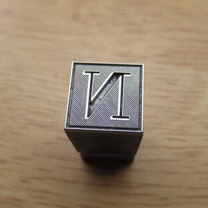 Può includere: Un timbro metallico quadrato con una lettera N incavata attraversata da una linea diagonale. La superficie del timbro presenta una trama finemente incisa in diagonale attorno alla lettera, il tutto racchiuso da un sottile bordo argentato in rilievo.