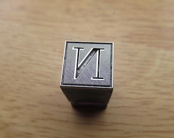 May include: A square metal stamp featuring a recessed letter N with a diagonal line through it. The face of the stamp has a fine, diagonal etched texture surrounding the letter, and the entire square is framed by a thin, raised silver-colored border.
