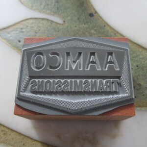 May include: A gray metal stamp with the text "OMAA TRANSMISSIONS" in a hexagonal shape. The stamp is mounted on a wooden block.