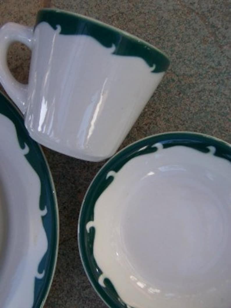 May include: White ceramic teacup and saucer set with a dark green scalloped border design. The teacup has a handle and the saucer is round. The set is clean and simple, suitable for a tea party or daily use.