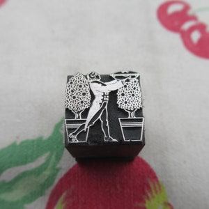 Może przedstawiać: Czarna gumowa pieczątka z wzorem przedstawiającym osobę niosącą tacę z dwoma doniczkami z drzewami. Osoba jest ubrana w garnitur i ma wąsy.