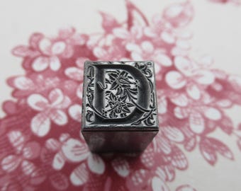 May include: A metal letterpress block with the letter "D" in a decorative floral design. The block is gray and has a slightly worn surface.