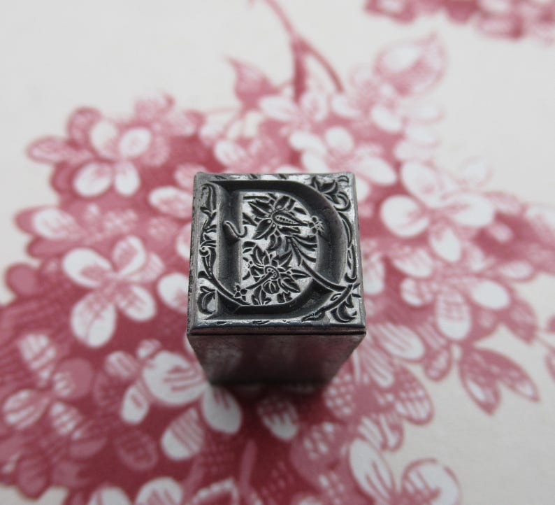 May include: A metal letterpress block with the letter "D" in a decorative floral design. The block is gray and has a slightly worn surface.