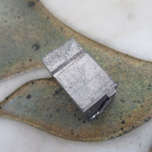 Op de afbeelding: Een close-up van een zilveren metalen blok met een rechthoekige vorm en een groef die door het midden loopt. Het blok heeft een ruw, getextureerd oppervlak.