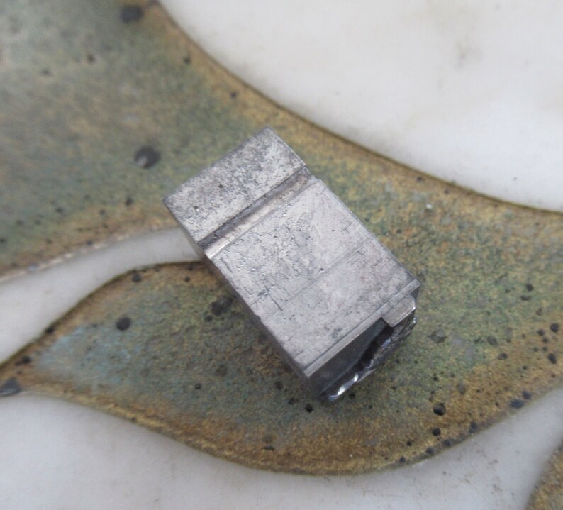 Op de afbeelding: Een close-up van een zilveren metalen blok met een rechthoekige vorm en een groef die door het midden loopt. Het blok heeft een ruw, getextureerd oppervlak.