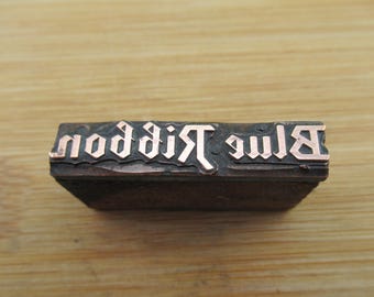 May include: A vintage metal printing block with the words "Blue Ribbon" in raised letters. The letters are filled with a copper-colored material.