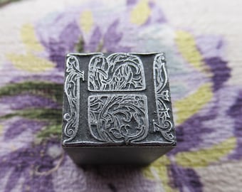 May include: A vintage metal letterpress printing block featuring the letter "H". The block is a dark gray color and has an ornate, floral design around the letter. The block is rectangular and has a solid base.