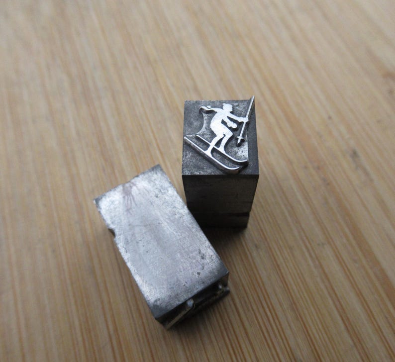 May include: Two vintage metal printing blocks. One block features a skier silhouette in silver. The other block is a rectangular shape with a worn, industrial appearance. The blocks are on a wooden surface.