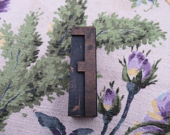 Può includere: Un blocco di stampa vintage in legno con la lettera "F". La lettera è marrone scuro con un centro nero, che mostra segni di usura. Il blocco è rettangolare e si trova su un tessuto a fantasia floreale con sfumature verdi e viola.