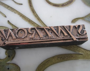 May include: A vintage metal printing block with the word "WYRON" in raised letters. The block is made of copper and has a dark brown wooden base. The text "REG TRADE MARK" is engraved on the base.