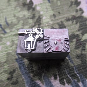 Op de afbeelding: Een vintage metalen drukblok met twee verschillende ontwerpen. Eén kant toont een gedetailleerde straatlantaarn met een brandende kaars, de andere een decoratief, mogelijk heraldisch embleem. Het blok is gemaakt van donker, verouderd metaal.