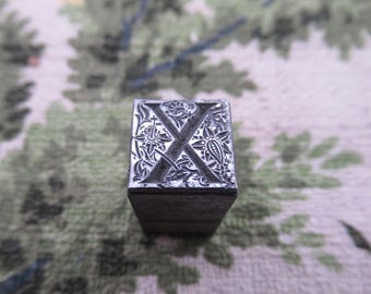 Könnte beinhalten: Ein Vintage-Metall-Letterpress-Druckblock mit dem Buchstaben "X". Der Block ist mit floralen und Paisley-Mustern verziert. Der Buchstabe ist in einer Serifenschrift. Der Block ist rechteckig und aus dunklem Metall.