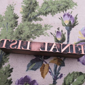 May include: A vintage copper letterpress block spells out "FINAL" in a serif font. The letters are mounted on a wooden base. The background is a floral patterned fabric with green, purple, and yellow hues.