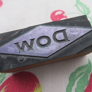 May include: A vintage printing block with the word "DOW" in a diamond shape. The block is made of wood and metal and has a purple background.