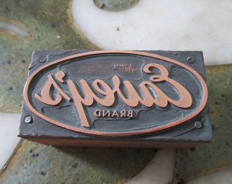 May include: A vintage metal printing block with a copper-coloured oval design. The oval features the text "Carey's Brand" in a stylised script font.