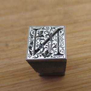 Può includere: Un timbro quadrato in stile vintage con le lettere "FM" in un design floreale nero decorativo. Le lettere sono su sfondo bianco, con una base in legno scuro. Il timbro è su una superficie di legno marrone chiaro.