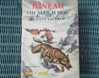 May include: A book cover with a white background and the title "Juneau The Sleigh Dog" by West Lathrop. The cover features an illustration of a dog pulling a sled with a person on it through a snowy landscape.