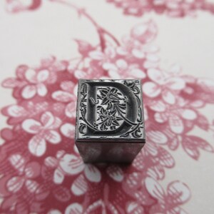 May include: A metal letterpress block with the letter "D" in a decorative font. The letter is surrounded by a floral design.