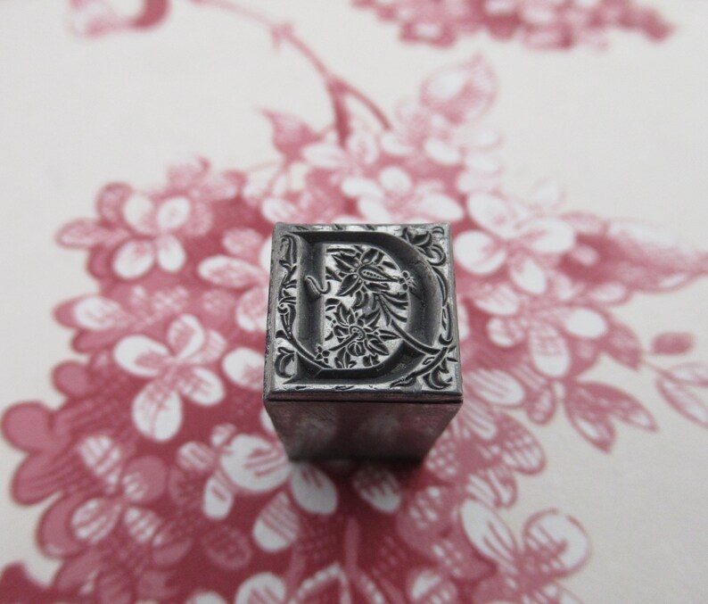 May include: A metal letterpress block with the letter "D" in a decorative font. The letter is surrounded by a floral design.