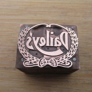 May include: A copper-colored printing block with the word "Daisies" in a decorative font, surrounded by a laurel wreath and a bow. The block is square and sits on a wooden surface.